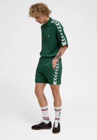 Zielona koszulka polo i szorty, wykonane z teksturowanej tkaniny, z kontrastującymi białymi paskami chevron na rękawach i bokach, w zestawieniu z paskowanymi skarpetkami i czarnymi butami.