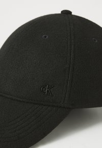 Black wool blend cap with a rounded crown, stitched detailing, and a glossy black cK logo on the front left side.