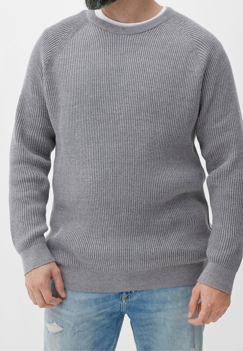 Mann, der einen grauen gerippten Strickpullover mit langen Ärmeln und hellblauen, zerrissenen Jeans trägt, vor einem schlichten Hintergrund steht.