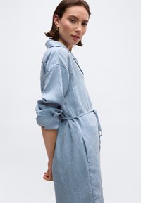 Γυναίκα που φοράει φόρεμα με μπλε και λευκές κάθετες ρίγες και μανίκια που έχουν γυριστεί, στέκεται πλαγίως μπροστά σε ένα απλό φωτεινό φόντο.