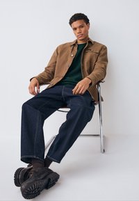 Brązowa koszula na guziki, zielony dzianinowy sweter, ciemne dżinsy i brązowe buty na krześle na białym tle. Zauważalne szczegóły szwów.