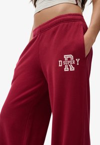 Bordeaux kleurige sweatpants van zachte stof, met een zijzak en een witte geborduurde logo op de dij. Elastische tailleband voor comfort.