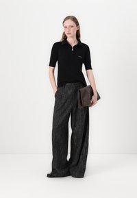 Polo shirt negru cu pliuri și guler cu fermoar, asortat cu pantaloni gri terciosi, largi. Modelul ține o poșetă maro. Design simplu, modern.