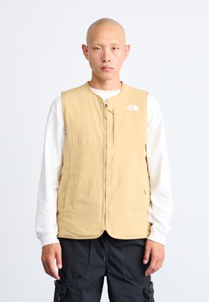 Light beige sleeveless vest made of soft fabric, featuring a front zipper and logo on the left chest, worn over a white long-sleeve shirt.