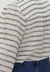 Lehká blůza v bílé barvě s černými pruhy. Texturovaná tkanina má vlnité linie a malé kulaté akcenty. Volný střih s nařasenými rukávy.