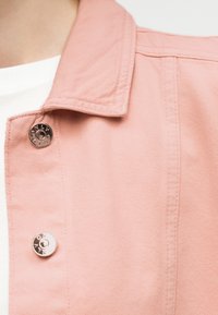 Κοντινό πλάνο ατόμου που φοράει ροζ τζιν μπουφάν με μεταλλικά κουμπιά πάνω από λευκό πουκάμισο.