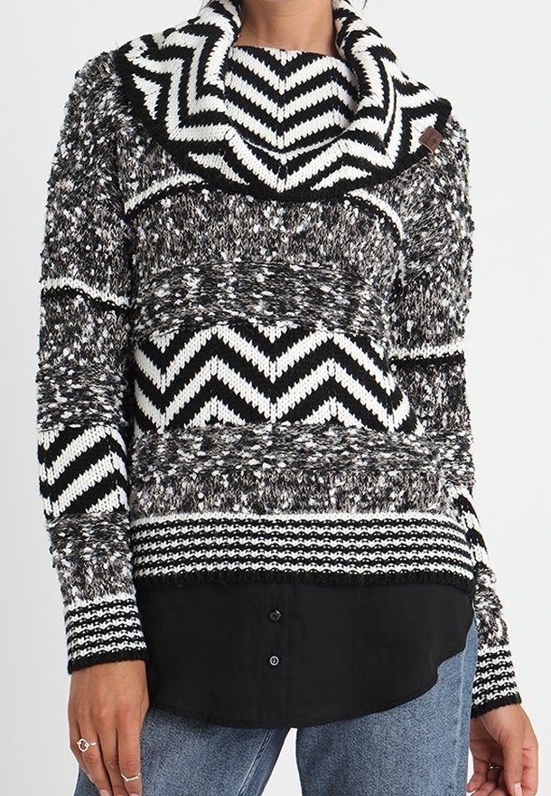 Pull noir et blanc tricoté avec des motifs en chevron et tachetés, grand col roulé, porté par-dessus une chemise noire boutonnée et un jean bleu.