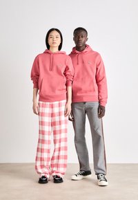 Δύο μοντέλα στέκονται δίπλα-δίπλα. Το ένα φοράει ροζ φούτερ και καρό παντελόνι, ενώ το άλλο φοράει ροζ φούτερ και γκρι τζιν με πλαϊνές λωρίδες.