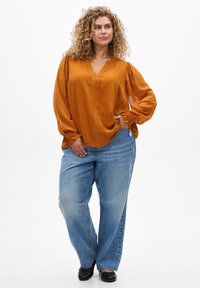 Senfgelb Bluse mit langen Puffärmeln, V-Ausschnitt und strukturierten Bündchen, kombiniert mit lockeren blauen Jeans und schwarzen Schuhen.