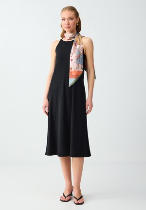 Donna che indossa un abito midi nero senza maniche, sandali neri con tacco, orecchini a cerchio dorati e una sciarpa colorata con fantasia annodata intorno al collo.