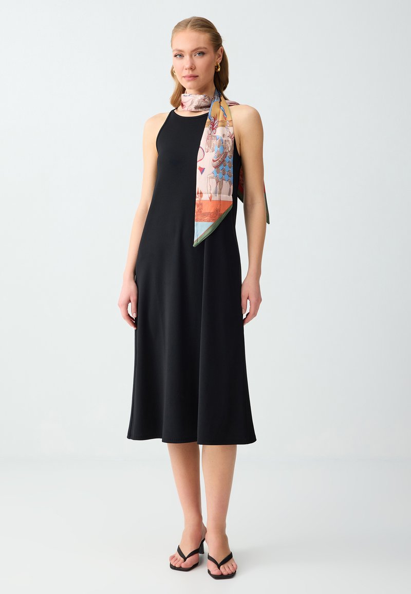 Femme portant une robe midi noire sans manches, des sandales noires à talons, des boucles d'oreilles créoles dorées et une écharpe colorée à motifs nouée autour du cou.