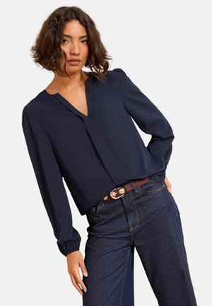 Jūras zila, garām piedurknēm blūze ar V veida kakla izgriezumu un plecu drapējumu, kombinēta ar tumšiem džinsiem un brūnu jostu ar metāla akcents.