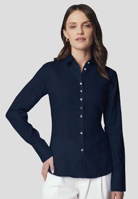 Granatowa koszula z długim rękawem i zapięciem na guziki o eleganckim kroju, z klasycznym kołnierzykiem i białymi guzikami, w parze z jasnymi spodniami. Prosta faktura materiału.