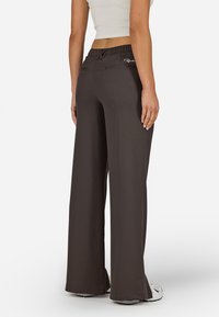 Kalhoty s širokými nohavicemi v tmavě hnědé barvě, s pružným pasem, dvěma zadními kapsami a hladkou texturou. Na nohavici není viditelné žádné logo.