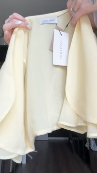 Blusa leve e fluida, de um amarelo pálido, com mangas curtas. Apresenta uma bainha arredondada, textura suave e etiquetas presas com cordões brancos.