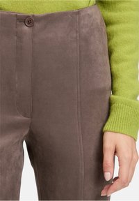 Close-up van bruine suède broek met knoopsluiting, gedragen met een groene gebreide trui, waarbij de hand van een vrouw naast de broek rust.