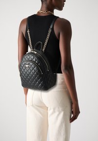 Sac à dos en cuir matelassé noir avec quincaillerie argentée, fermetures éclair doubles et une chaîne sur les bretelles, porté par une personne en tenue décontractée.