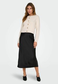 Cardigan corto avorio con bottoni frontali e due tasche, abbinato a una gonna midi nera in satin e scarpe slip-on nere.