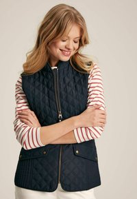 Tengerészkék steppelt mellény magas gallérral és cipzárral az elején, arany színű díszítéssel és oldalsó zsebekkel, vörös és fehér csíkos hosszú ujjú póló fölött viselve.