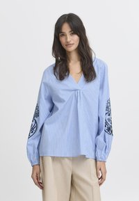 Bluza prugasta plava i bijela s dugim rukavima koja sadrži crnu cvjetnu vez na manžetama. Dizajn s V-izrezom, ležeran kroj.