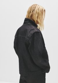 Svart denimjacka med högt krage, synliga sömmar och framfickor. Tyget har en lätt lyster och en strukturerad silhuett.