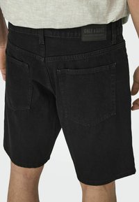 Pantalones cortos de mezclilla negros con corte recto, con dos bolsillos traseros y una etiqueta en la cinturilla, mostrados desde la parte trasera.
