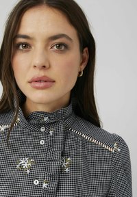 Blouse à carreaux noir et blanc avec un col à volants, des boutons sur le devant, des détails floraux brodés et des accents rayés sur les épaules.