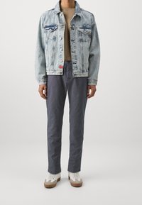 Kurtka dżinsowa w jasnoniebieskim kolorze, dwie kieszenie na piersi, beżowy sweter, szare spodnie oraz białe trampki z brązowymi akcentami.