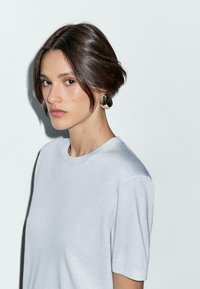 Giovane donna con capelli scuri della lunghezza delle spalle e una maglietta grigia chiara, che guarda dolcemente verso la fotocamera su uno sfondo bianco semplice.