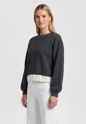 Woman with straight blonde hair wearing a dark gray long-sleeve sweater over a white shirt and white pants, standing against a plain background.