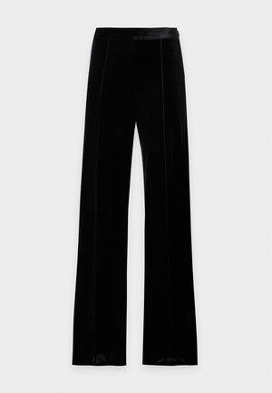 Femme debout portant une chemise en soie turquoise avec des imprimés de calèches et un pantalon large noir, sur un fond blanc uni.