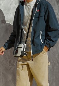 Marinblå zip-up jacka med vita accenter, bärs över en vit t-shirt, tillsammans med beiga byxor och en grå och svart axelväska.