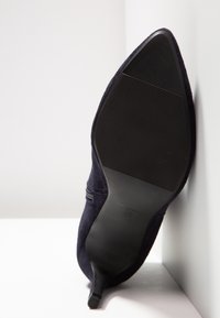 Chaussure à talon haut en daim noir unique, affichée de côté, montrant la semelle texturée et l'extrémité pointue sur un fond blanc en angle.