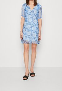 Robe florale bleu clair avec de courtes manches transparentes, décolleté en V et ourlet au-dessus du genou. Associée à des sandales noires à bout ouvert avec des brides torsadées.