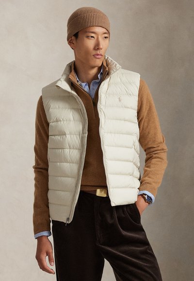 Lysebeige, oppustet vest med høj krave, båret over en brun sweater. Bemærkelsesværdig ribstrikket brun hue og mørke kordfløjlsbukser med lynlås.