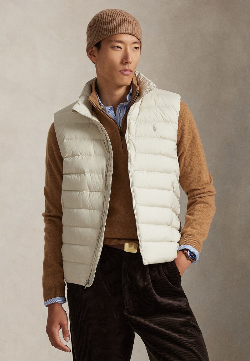 Ljusbeige puffig väst med hög krage, bärs över en brun tröja. Framträdande ribbad brun hatt och mörka corduroybyxor med dragkedja.