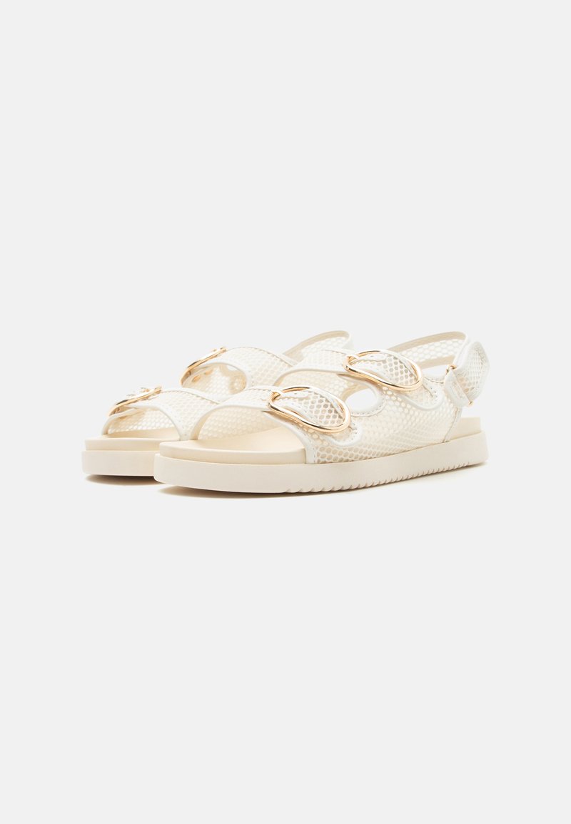 Sandalias blancas de punta abierta con tiras de malla, hebillas doradas y suelas planas, mostradas en un ángulo ligero sobre un fondo neutro.