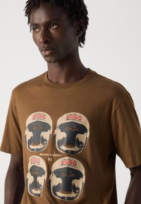 Junger Mann in einem braunen T-Shirt mit vier Druckmotiven von Boxhelmen, die mit "HUGO" beschriftet sind, sowie dem Text "BOXING ACADEMY 93" auf der Vorderseite.