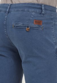 Blue denim trousers featuring a back pocket with a button closure and a brown leather brand patch. The fabric has a textured surface.