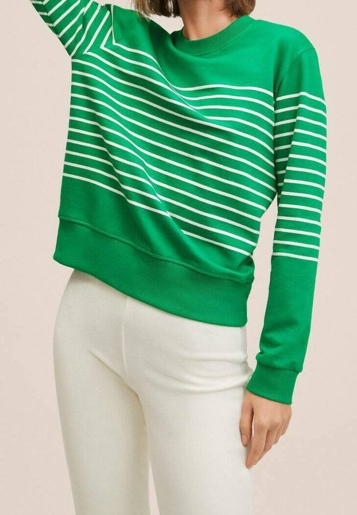 Femme portant un sweat-shirt vert à rayures blanches horizontales et un pantalon ajusté blanc, posant avec un bras levé.