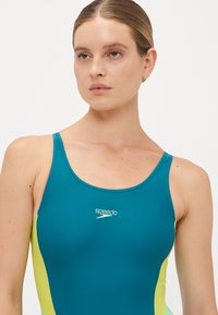 Kvinne med lys hud og brunt hår iført en turkis og gul Speedo badedrakt, som ser litt til siden mot en enkel hvit bakgrunn.