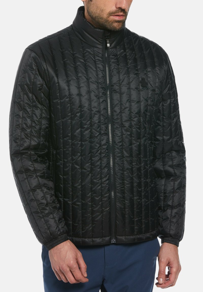 Mann trägt eine schwarze gesteppten Reißverschlussjacke mit Stehkragen und elastischen Bündchen, kombiniert mit dunkelblauen Hosen, vor einem weißen Hintergrund.