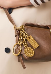 Bruine leren handtas met gouden ketting en drie gestructureerde gouden charme's, vastgehouden door een persoon in een beige jas met zwarte knopen.