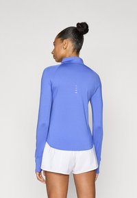 Αθλητικό τοπ με μακριά μανίκια σε ζωντανό μπλε χρώμα, με ψηλό γιακά και διακριτικές λεπτομέρειες στην πλάτη. Λεπτό, ελαφρύ ύφασμα με εφαρμοστή γραμμή.