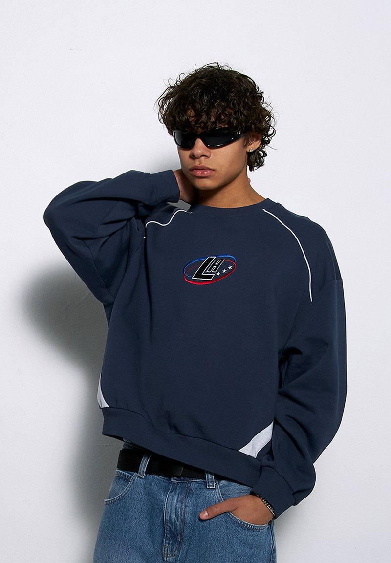 Granatowa bluza z białymi akcentami, okrągłym dekoltem i haftowanym logo na piersi. Noszona z jasnoniebieskimi dżinsami i okularami przeciwsłonecznymi.