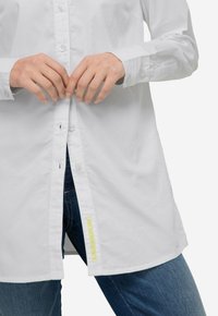 Camicia bianca con bottoni e maniche lunghe, in tessuto di cotone, dal taglio dritto, con un'etichetta gialla sul lato, indossata sopra un paio di jeans di denim.
