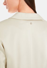 Beige collar shirt with a smooth texture, featuring a back button detail and a vent at the center, emphasizing a minimalist design.
