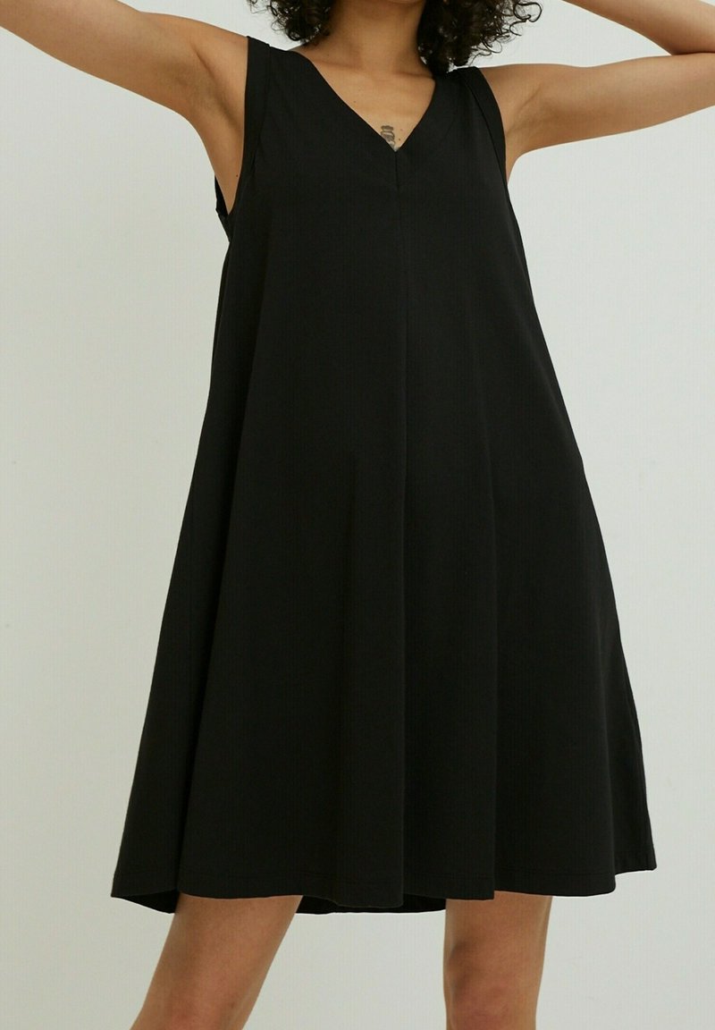 Femme portant une robe noire ample sans manches avec un col en V, debout les bras levés devant un fond uni clair.