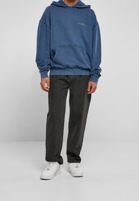 Felpa in denim blu con tasca frontale e cappuccio regolabile con cordino, abbinata a jeans neri larghi e sneakers bianche.