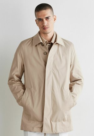 Man wearing a beige buttoned coat with hands in pockets, standing against a plain light background.